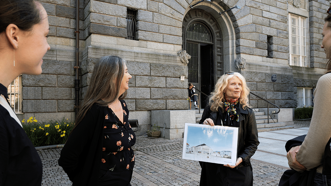 En kvinne står foran en eldre bygning i granitt. Til en liten gruppe viser hun en tegning. 