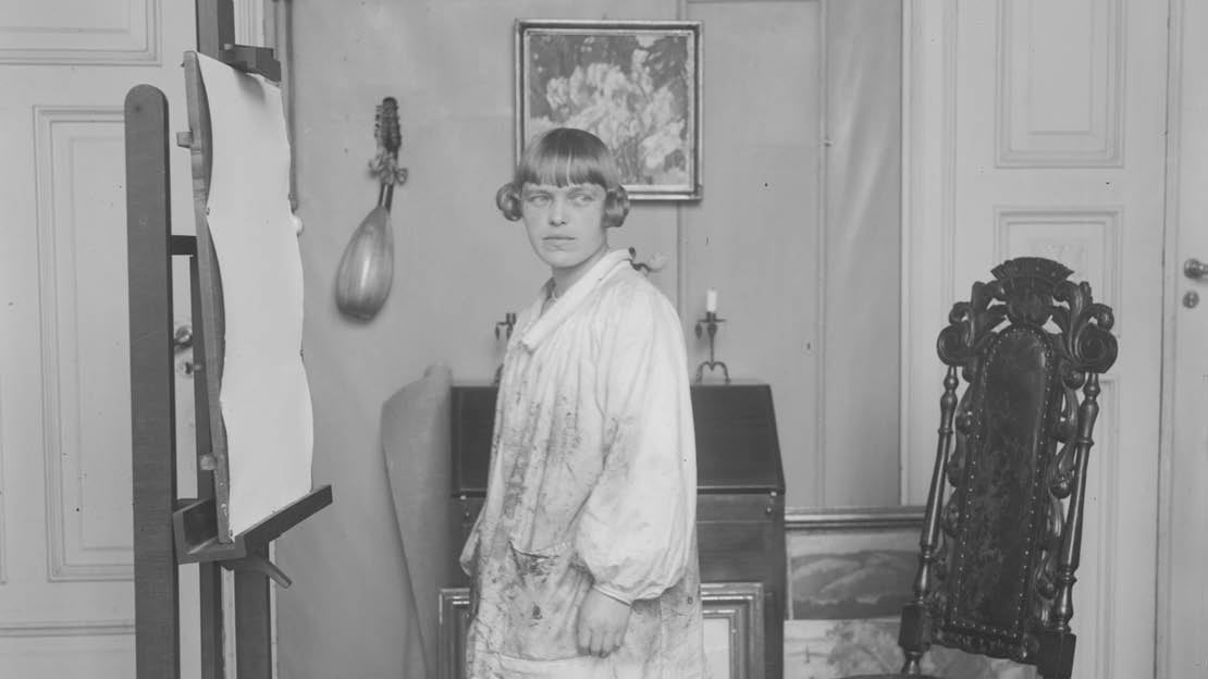 Svart-hvitt fotografi som viser kunstneren stående i atelieret med staffeli foran seg og bilder på veggen bak.
