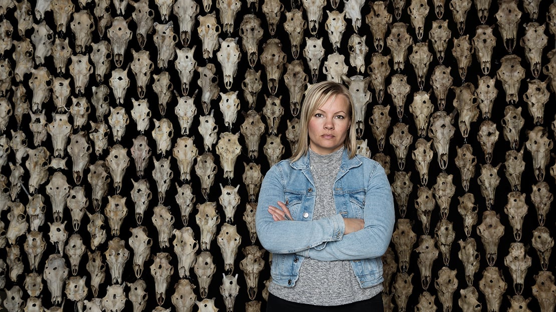 Máret Ánne Sara her foran sitt eget verk «Pile o’Sápmi Supreme» Kvinne med lyst hår, kledd i olajakke står med armene i kors foran et hengende teppe av reinsdyrhodeskaller