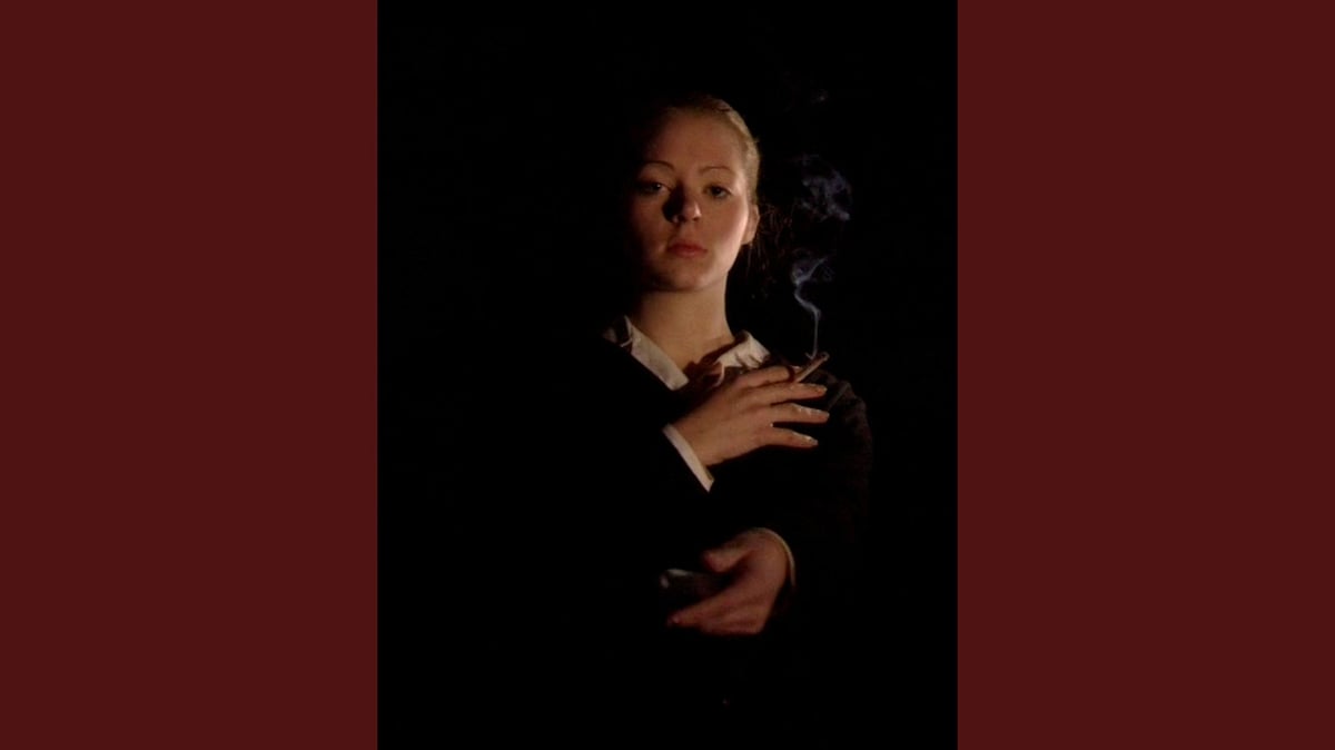 A K Dolven, «portrait with cigarette», 2000 Bildet viser en kvinne som ser mot oss, hun holder en sigarett i den ene hånden og en fjernkontroll i den andre.