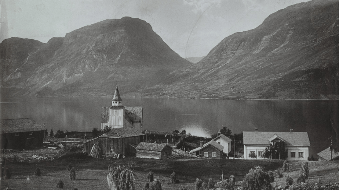 Gammelt svart-hvitt foto av landskap med fjell, innsjø, stavkirke og gårdsbygninger; i forgrunnen høystakker.