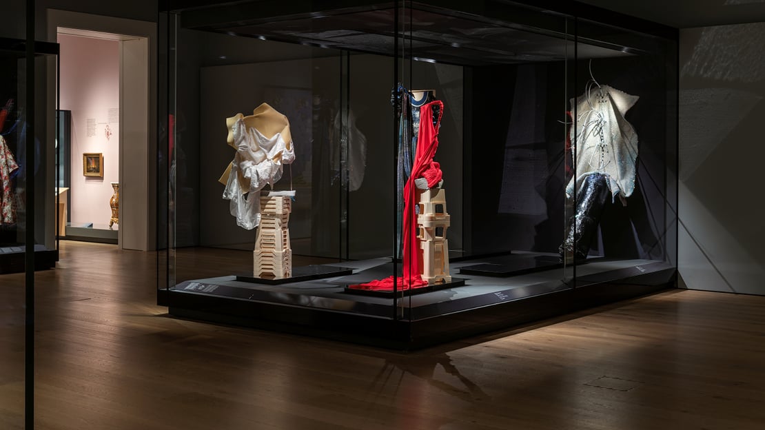 I et mørklagt museumsrom er fire antrekk som ligner en blanding av klær og skulptur, utstilt i en monter. 
