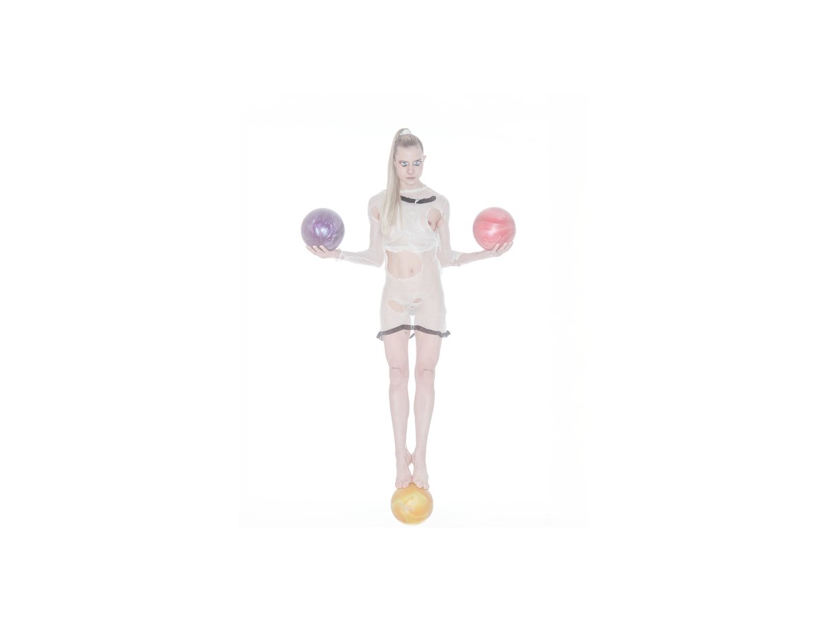 A person balances on a yellow ball while holding a purple and a pink ball, wearing a transparent outfit.