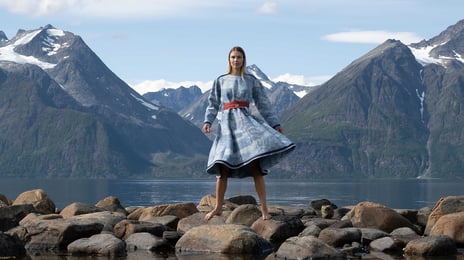 En kvinne står foran et fjell, kledd i en kjole med mønster og farger fra fjellet. I forgrunnen ser man henne i et vannspeil.