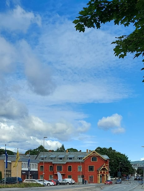 Sandsgården på Gjøvik, sommeren 2020 Fotografi. Blå himmel med skyer, grener med grønne blader i forgrunnen, nederst en rød bygning med gule vinduskarmer og detaljer