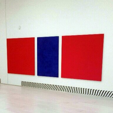 Three square canvases on a white wall. Two painted all red, the middle one painted all blue.