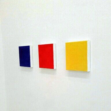 Three square canvases on a row on a white wall. Left to right: one blue, one red, one yellow canvas.
