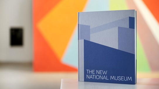«Det nye Nasjonalmuseet» av Orfeus forlag/Nasjonalmuseet. Redaktør/hovedforfatter: Audun Vinger. Formgitt av Kørner & Ludvigsen Nærbilde av en blå bok med stilisert bygning og teksten "the new national museum"