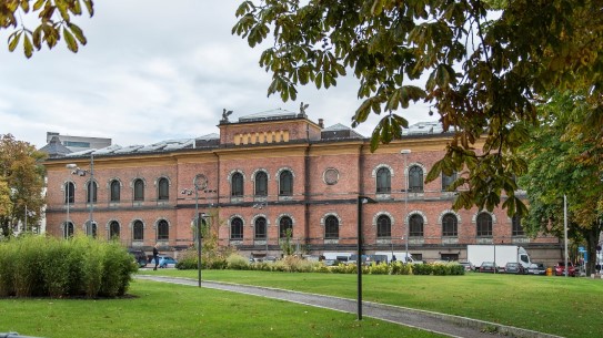 Blir det ny inngang til Nasjonalgalleriet og uteservering på Tullinløkka? Gammel teglsteinsbygning. Park med plen, trær og busker i forgrunnen. Biler parkert langsmed fasaden.