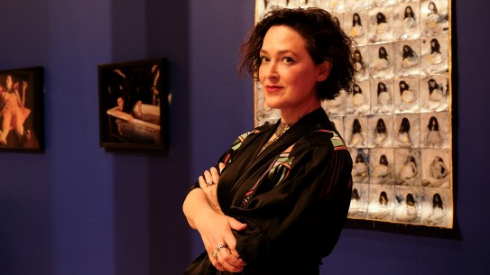 Hettie Judah. Portrait of curator and writer Hettie Judah, standing with arms crossed in front of an artwork on a purple wall.
