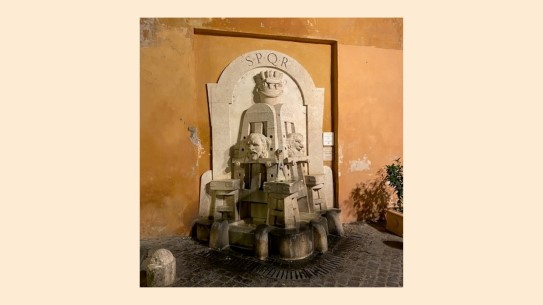 «Fontana degli Artisti», Roma. Fotoet viser en utendørs situasjon. Mot en okergul, kalket husvegg er en skulpturell fontene med en flat portalform mot veggen