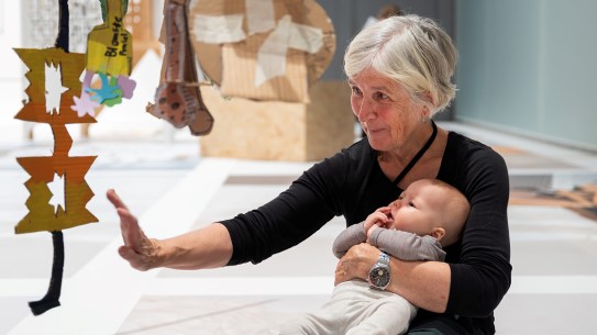 Alt.tekst: En kvinne har en baby på fanget. De ser mot en installasjon fra utstillingen Hånd og Maskin.