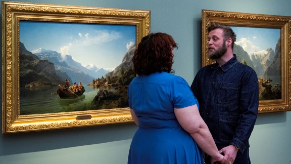 En dame og en mann står vendt mot hverandre og holder hender foran maleriet «Brudeferd i Hardanger», mens mannen synger. 