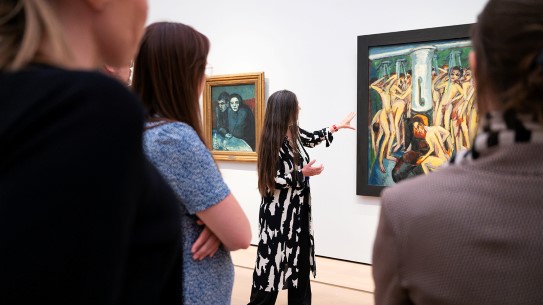 En formidler peker mot et kunstverk. I forgrunnen står man de som hører på.