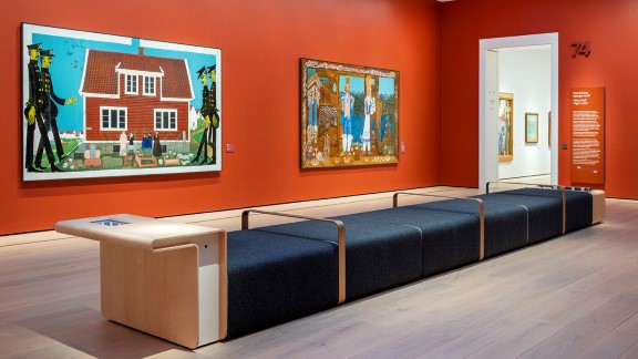 Et rom med orange vegger, hvor det henger to store malerier. I midten av rommet er det en lang sittebenk. 