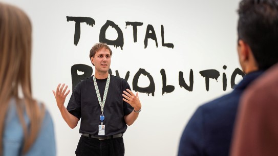 En mannlig omviser står foran verket "Total revolution". Kunstverket er synlig på veggen bak ham.