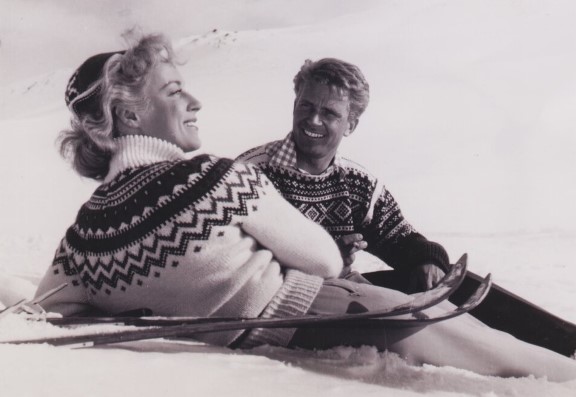 To blide mennesker ligger i skibakken i sola med strikkagensere på