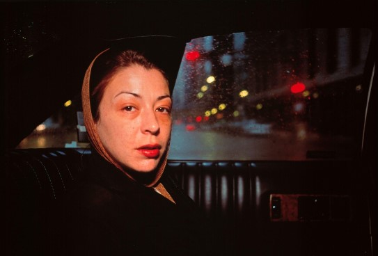 Nan Goldin, «Kathleen in the Taxi to Greer's Funeral. Chicago»¸ 1996. En kvinne ser mot oss fra baksetet i en taxi, i en mørk bygate, med trafikklys i bakgrunnen
