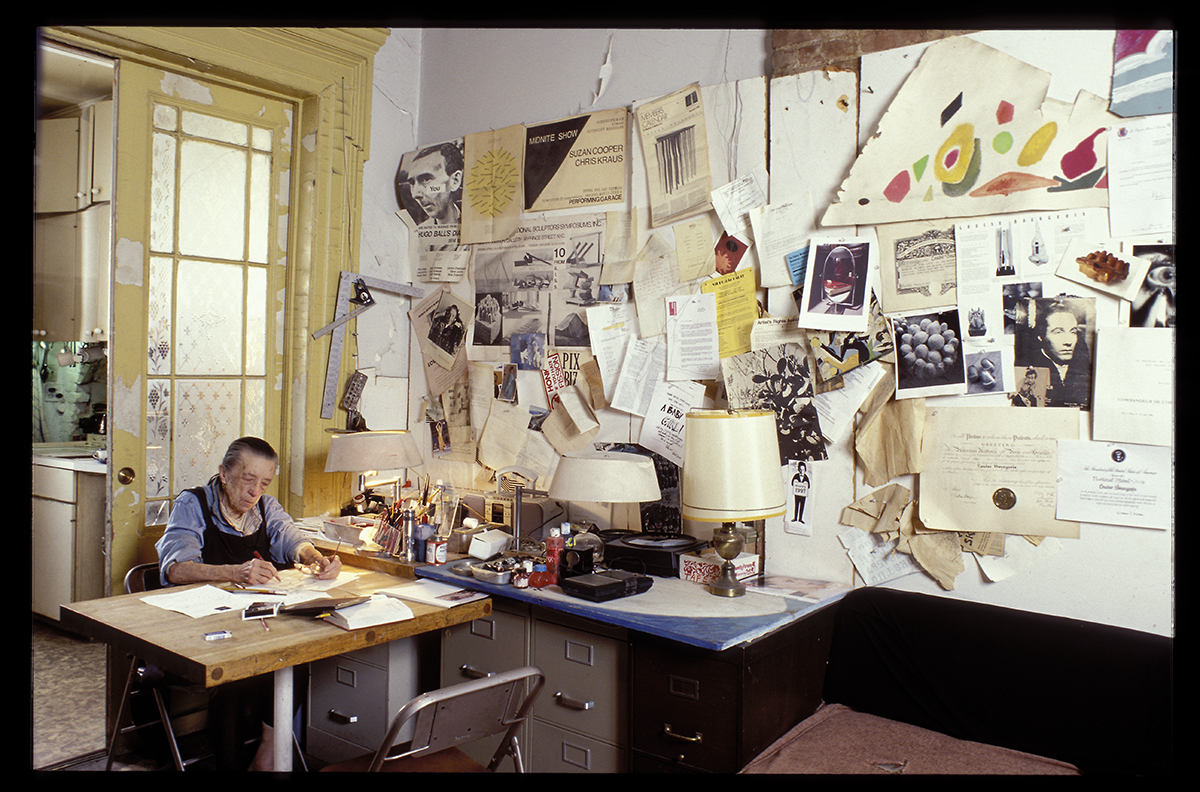 Older lady sitting down at a desk in front of a wall covered with different things