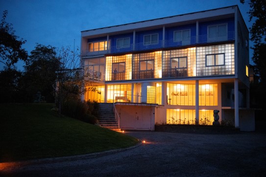 Hvitt hus med flatt tak i mørke, opplyst innenfra gjennom vinduer og glassvegg, Mørk blå himmel over huset og hagen.
